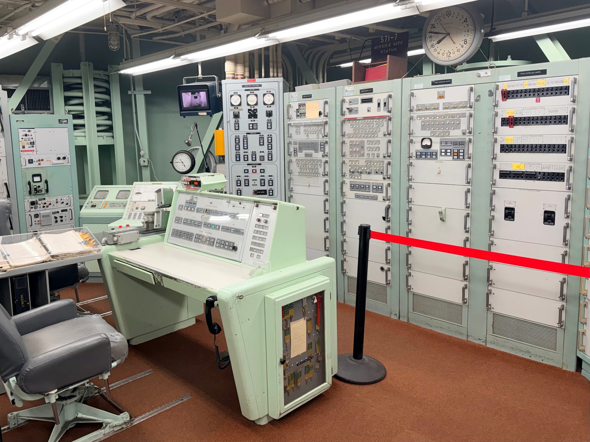 A photo showing the view from the right side of the control room. A desk sits in the middle of the frame, with electronic equipment cabinets in front of the desk.
