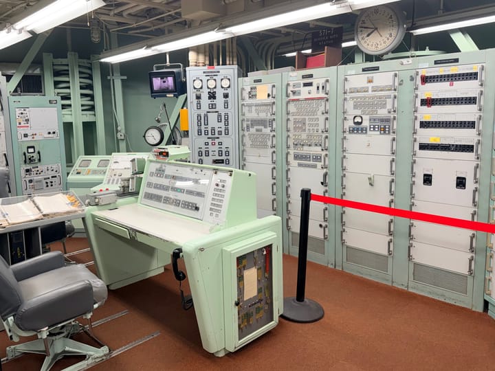 A photo showing the view from the right side of the control room. A desk sits in the middle of the frame, with electronic equipment cabinets in front of the desk.